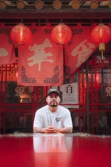 A man wearing a white shirt and a black cap sits at a table inside a traditional Chinese restaurant. Red lanterns hang above, and the decor features Chinese characters and designs.