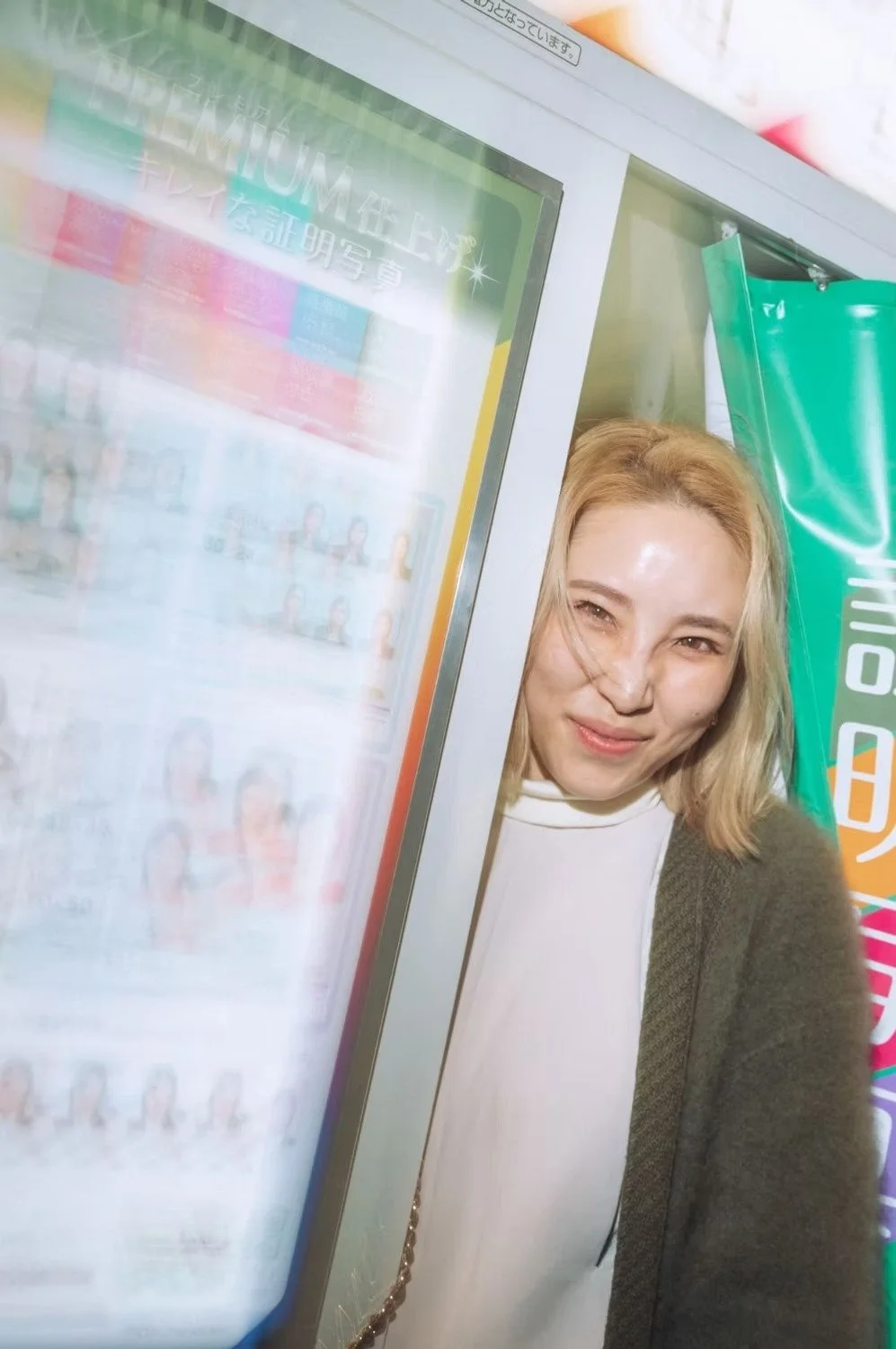 A young woman smiling and playfully peeking out from behind a vending machine on a colorful Tokyo street. Street photography by Deniz Demir.