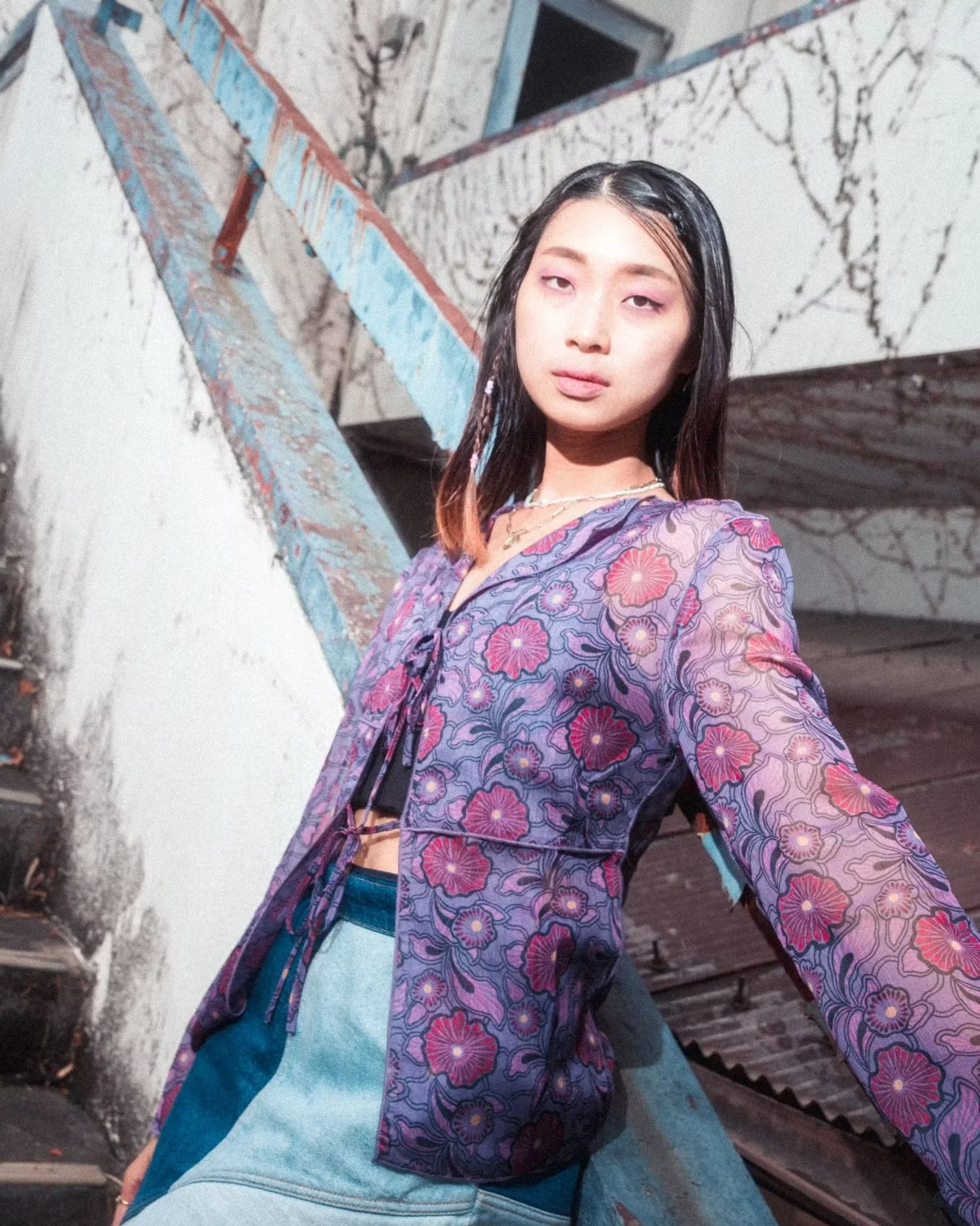 A young woman with wet hair wearing a purple floral top taking a selfie outdoors in front of a weathered wall with graffiti and a staircase.