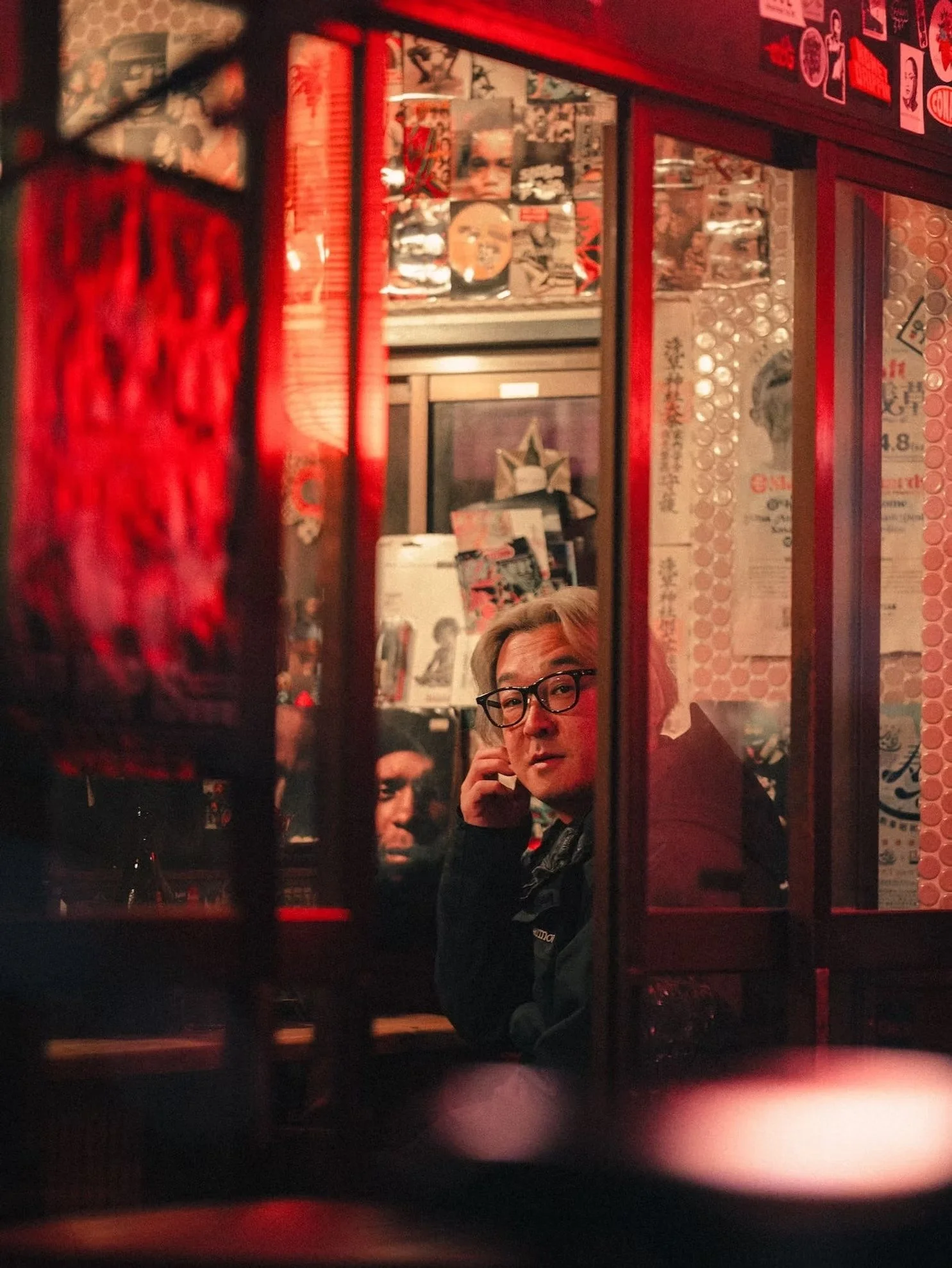 A man with blonde hair and glasses looking at a camera through a mirror, with colorful posters and decorations in the background, inside a dimly lit restaurant or bar with red lighting.