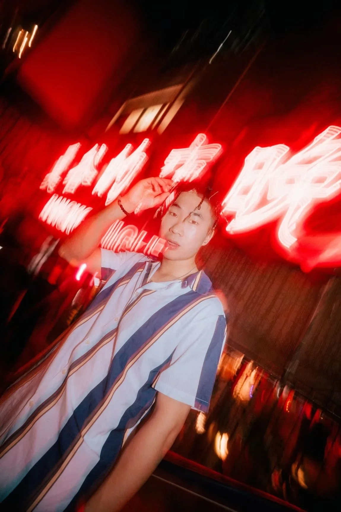 A young man in a striped shirt posing confidently under bright red neon lights in Tokyo at night. Street photography by Deniz Demir.