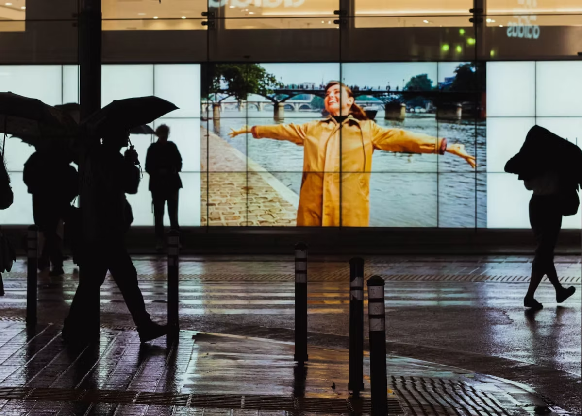 People walking in the rain with umbrellas in front of a large outdoor screen showing a woman in a yellow raincoat with arms outstretched near a river and bridge.
