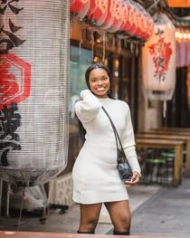 A woman in a white dress with a sleek black bag smiles while posing next to a traditional Japanese lantern outside a shop. The lantern is decorated with bold red and black Japanese characters.