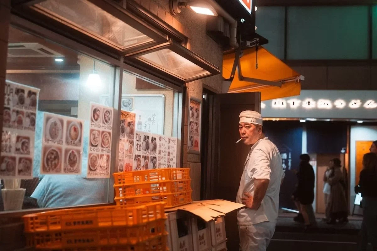 A man dressed as a chef standing outside a Japanese restaurant at night, smoking a cigarette. The restaurant window displays menu pictures and orange crates are stacked outside. Shibuya nights shot by Tokyo street photographer Deniz Demir.