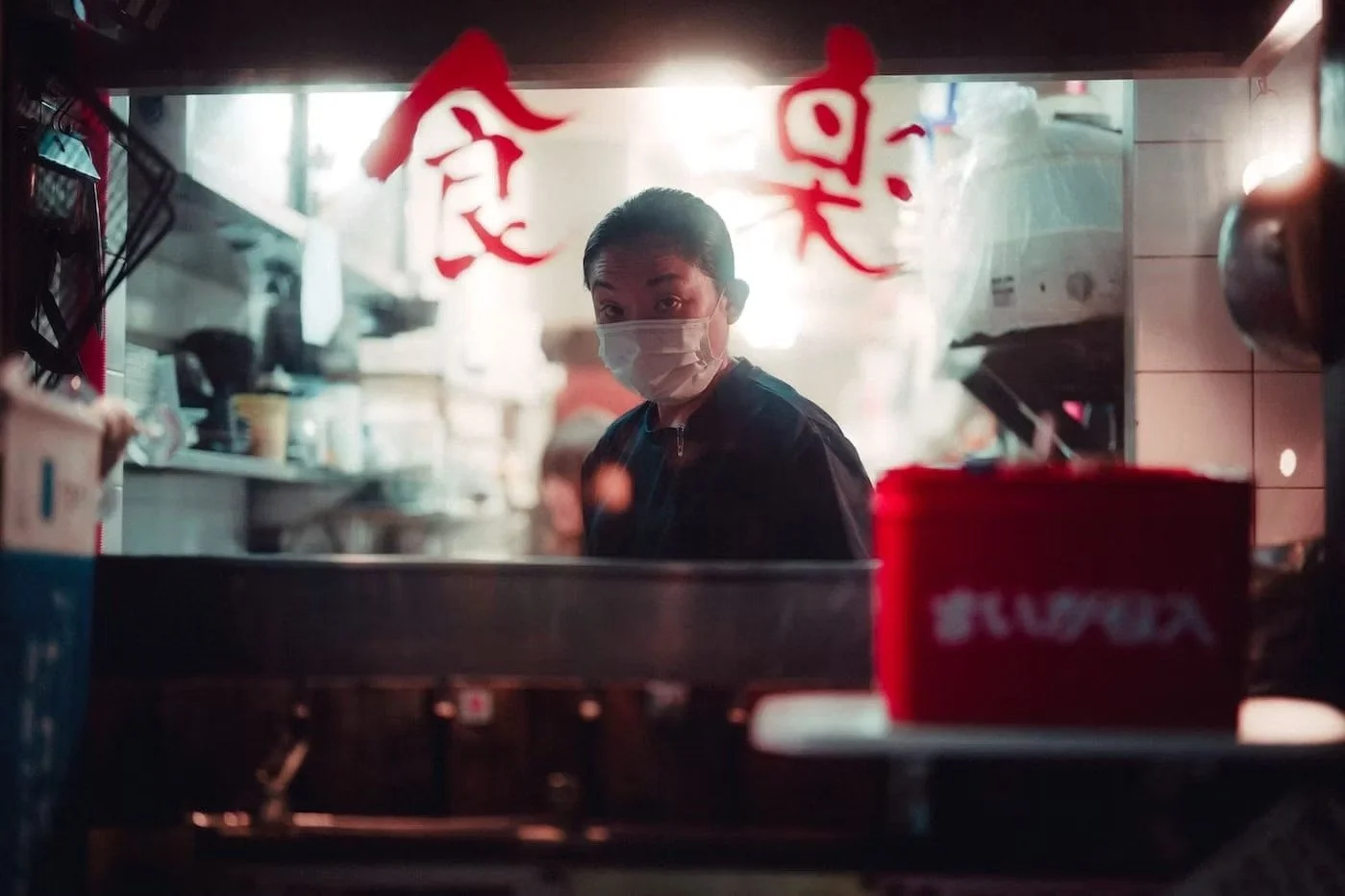 A person wearing a face mask looks through a restaurant window with red Chinese characters, kitchen equipment, and a red container visible inside. Shot in Ikebukuro, Tokyo.
