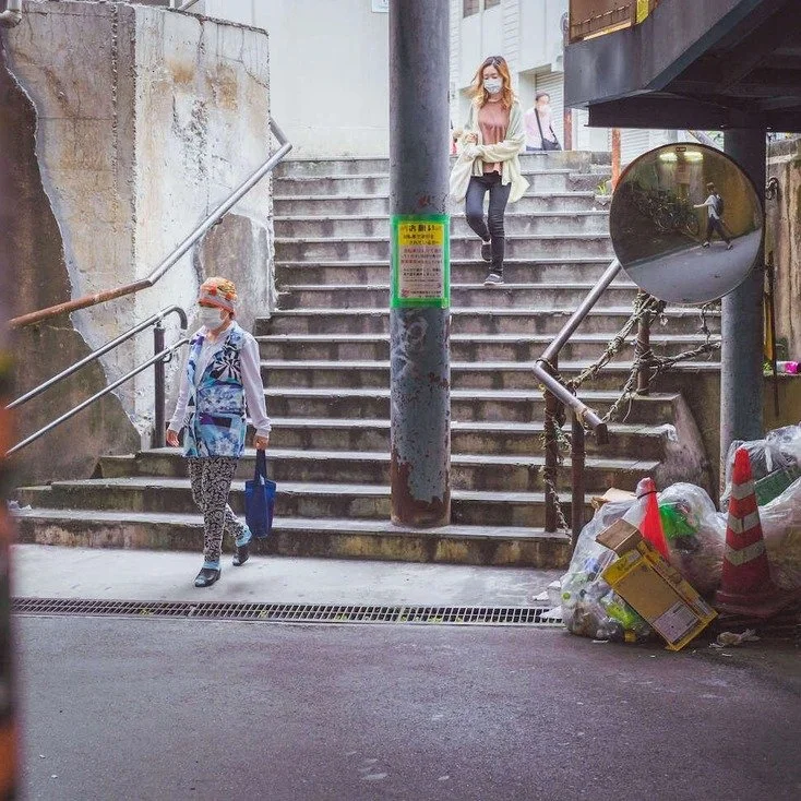 Two people walking up the stairs of an Osaka street, with trash bags and a sign visible, captured during slow travel.