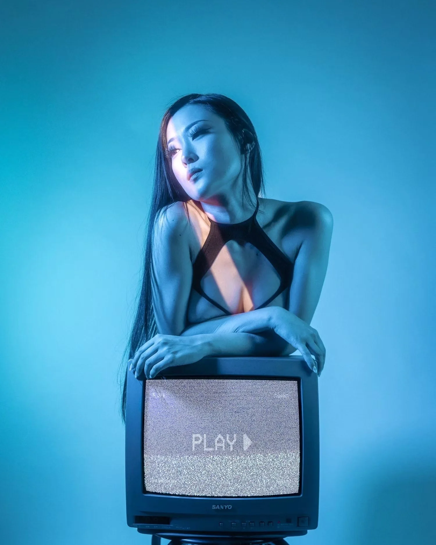 A woman with long dark hair sitting behind an old television set with a screen displaying the word 'PLAY' in white text on a gray background. The woman is looking to her left, wearing a sleeveless black top, and partially leaning on the TV with her a