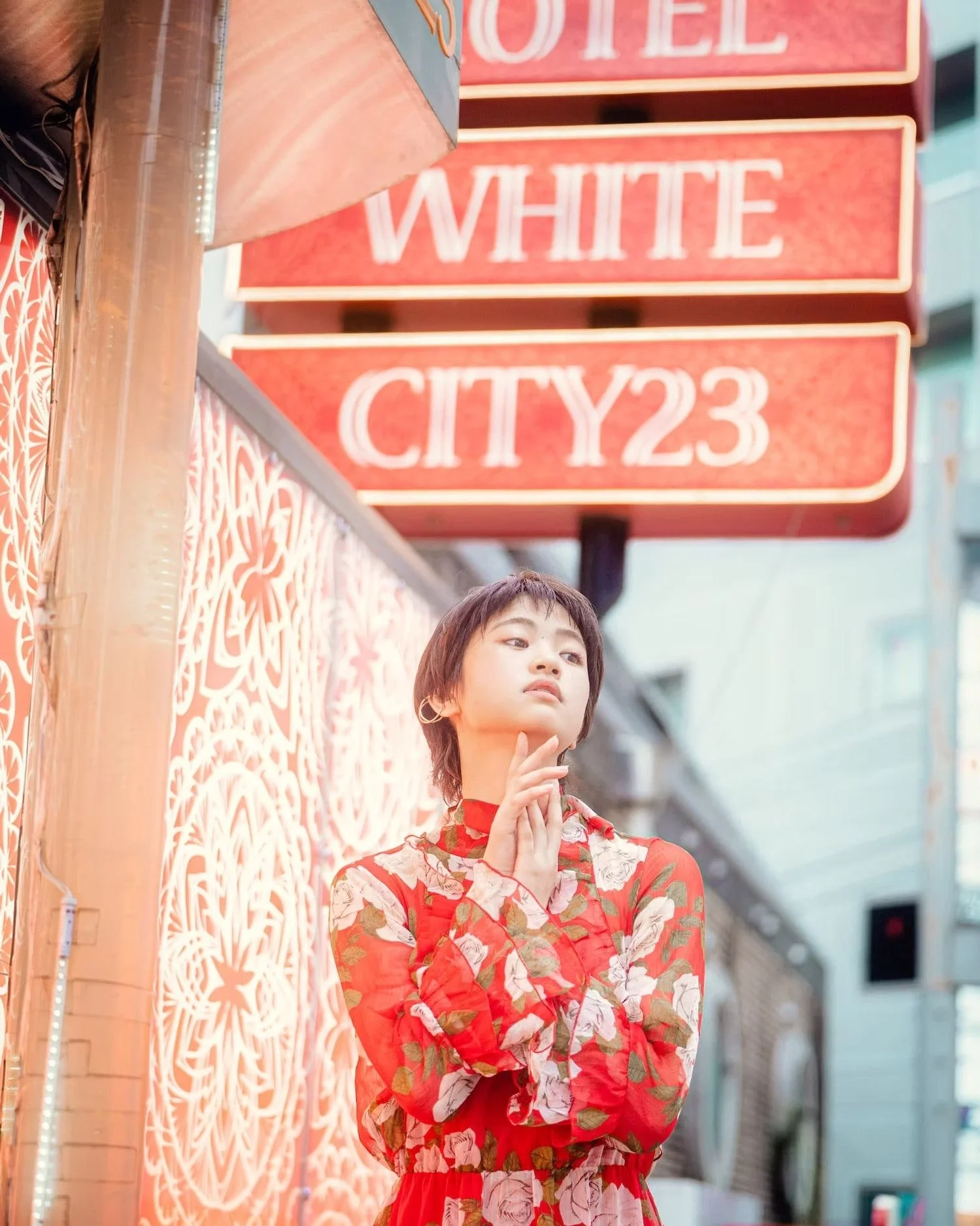A young woman with short brown hair and hoop earrings standing outdoors in front of a decorative wall, wearing a red floral dress. There is a red street sign above her with the white text 'WHITE CITY 23' and an orange wooden railing to her side.