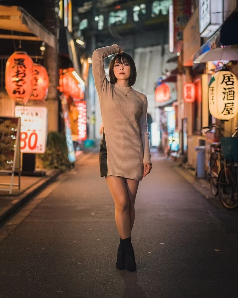 A woman standing in the middle of a lit street at night, surrounded by Japanese lanterns and signs, with her hand resting on her head and wearing a long sweater, shorts, and high socks.