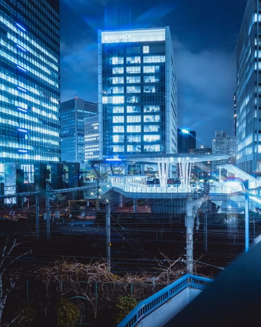 Night view of a modern city with illuminated skyscrapers and a footbridge over train tracks.