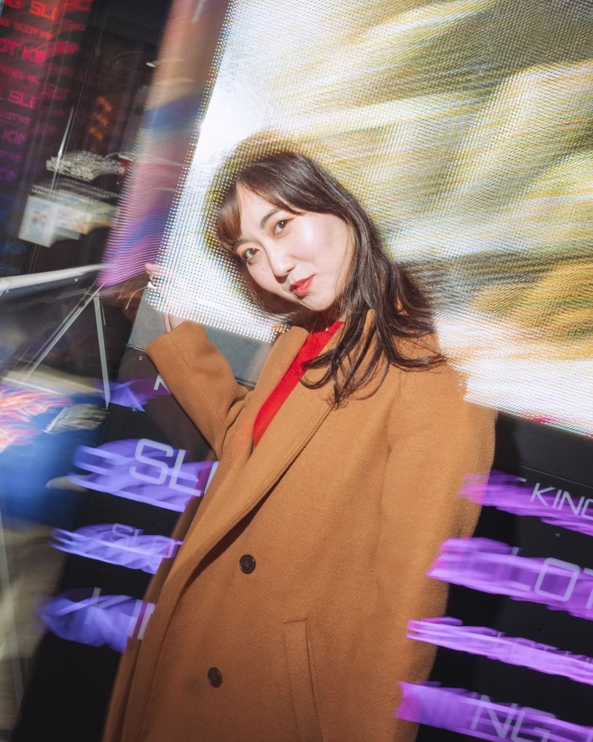 A woman stands confidently in front of a brightly lit backdrop with neon purple lights, creating a bold and dramatic look. The vibrant lighting contrasts with her outfit, adding depth and energy to the urban portrait.