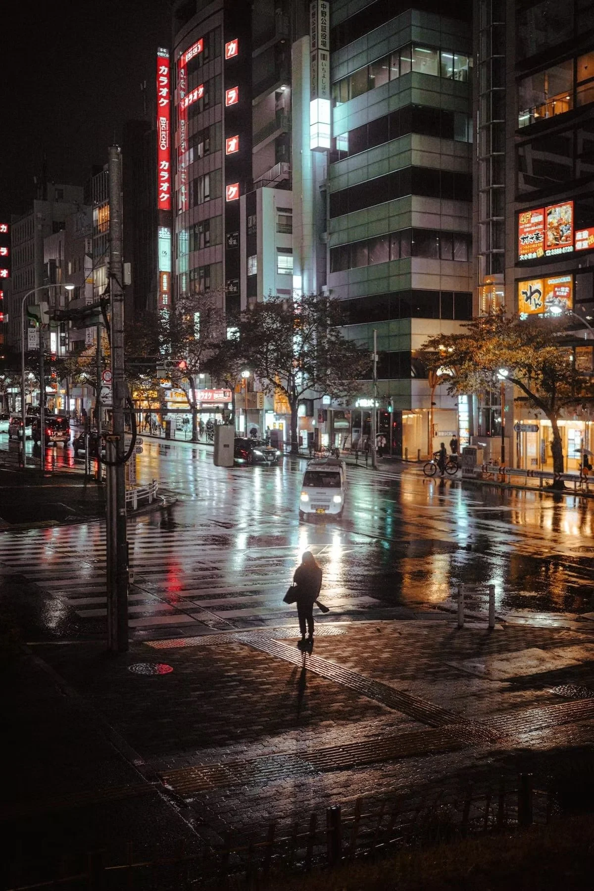 Nighttime city street scene in Nakano Tokyo with wet roads reflecting neon lights, a person crossing the street, cars, and buildings illuminated by signs and lights.