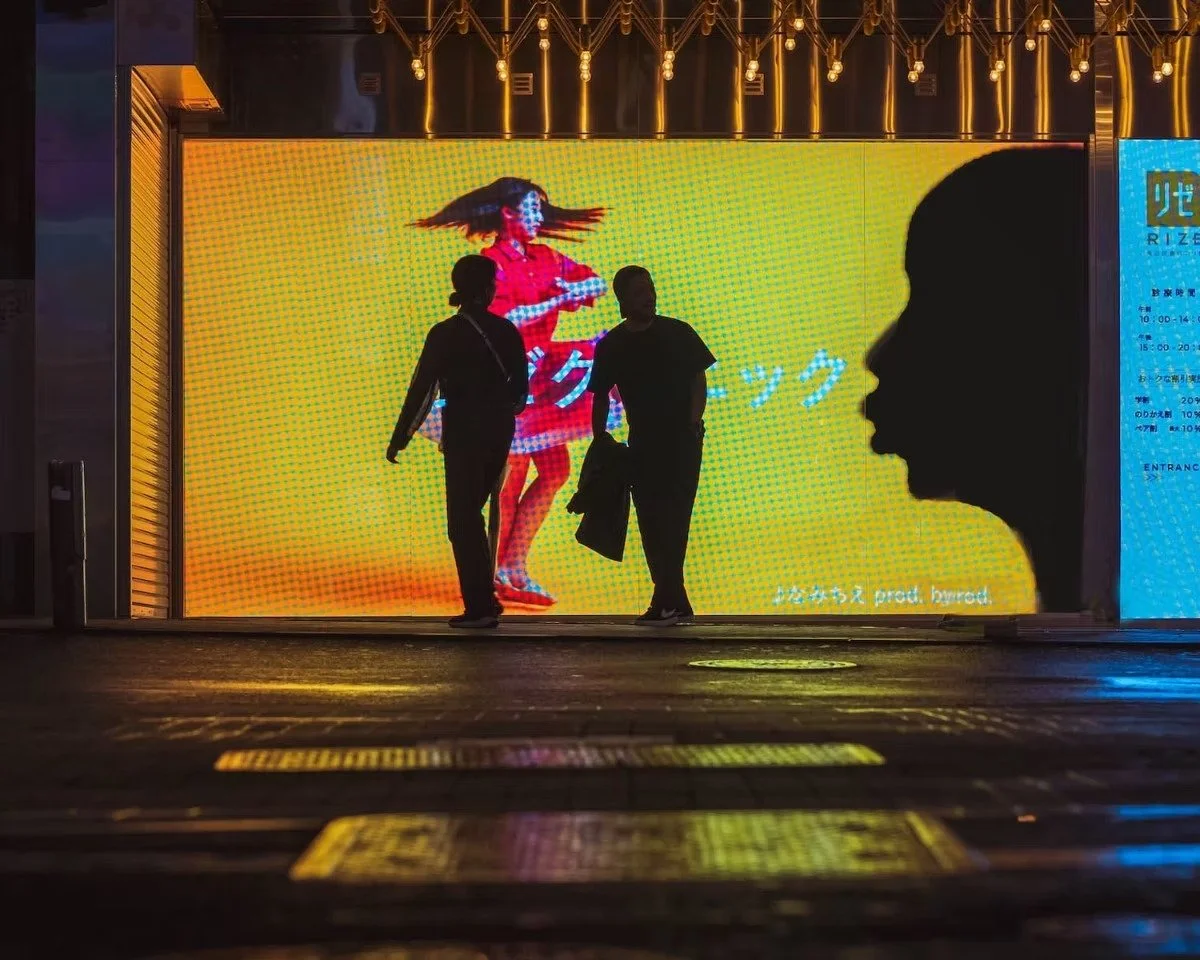 Silhouettes of two people walking in front of a brightly lit advertisement display with a colorful image of a woman and Japanese text in Shibuya.