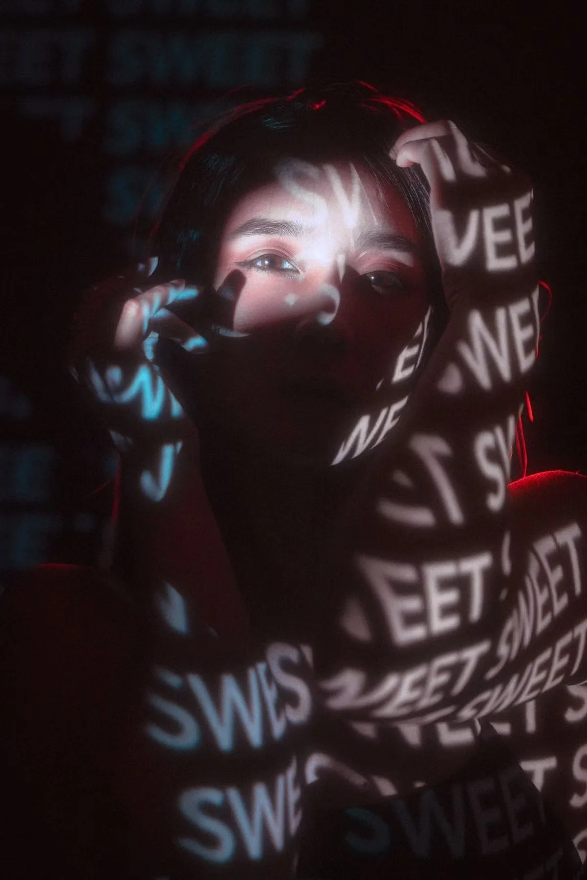 A person with short dark hair and light skin is holding up their hands near their face, partially covering their features. The person's face and hands are illuminated by a projection of the word 'SWEET' repeatedly displayed in white letters, creating