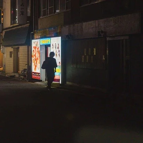 A lone figure walks past a brightly lit vending machine in a dark Tokyo alley, creating a mysterious and cinematic urban scene.