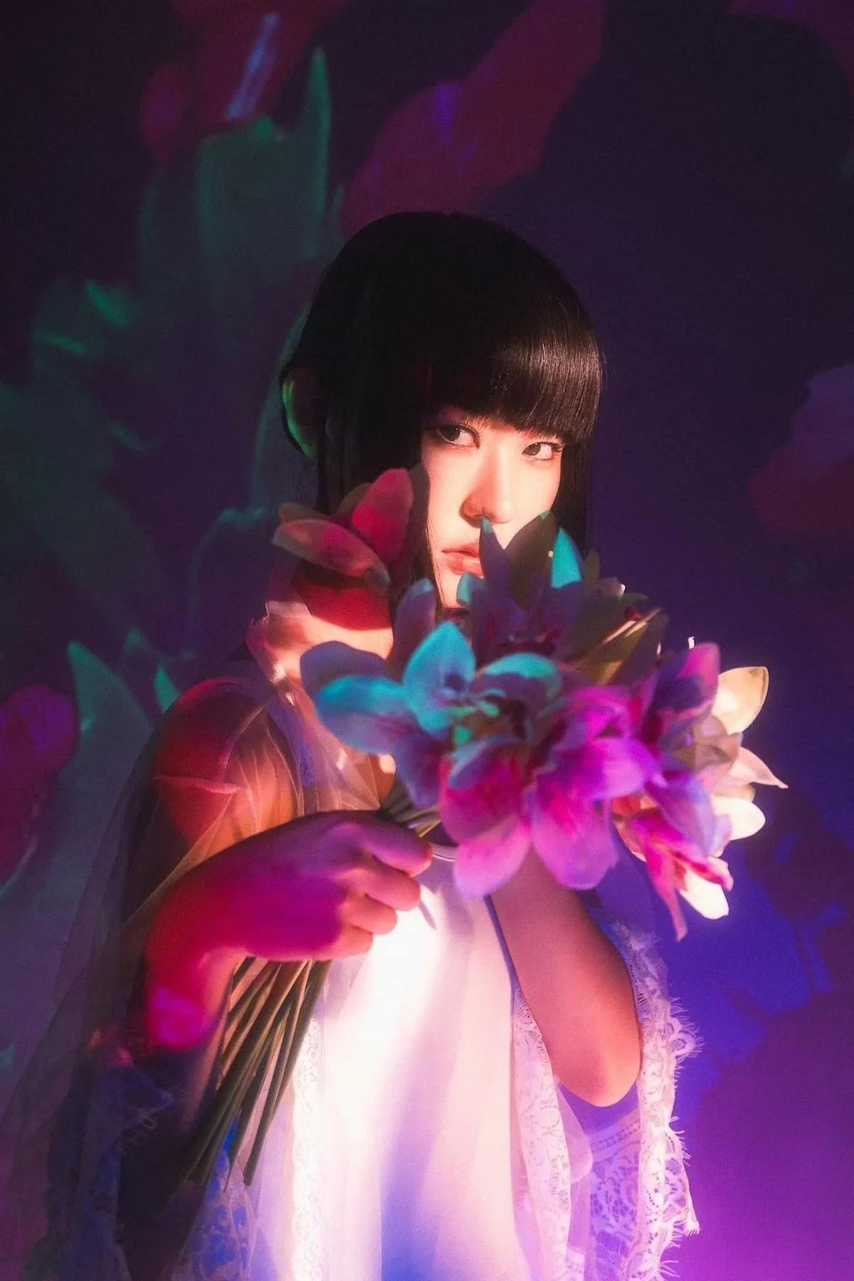 Studio portrait photography of a woman with black hair and bangs holding a colorful bouquet of flowers, illuminated by vibrant purple, blue, and pink lighting.