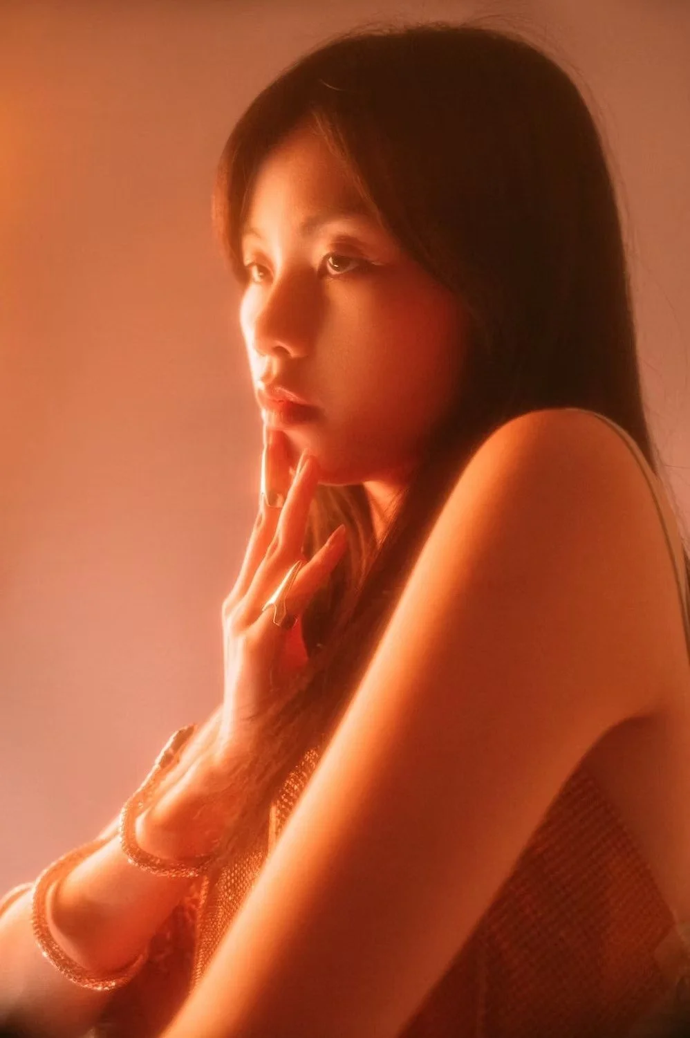 A close-up of a woman in soft orange lighting, with her hand on her chin in a thoughtful pose. The warm light highlights her profile, creating a contemplative and serene atmosphere.