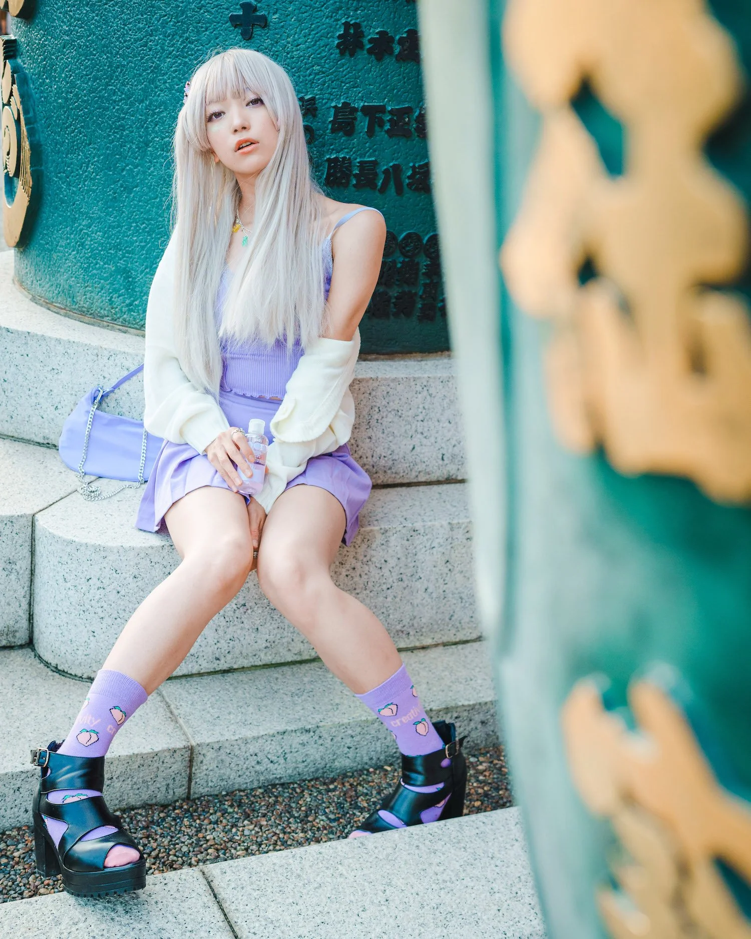 A young woman with long, platinum blonde hair is sitting on stone steps against a green wall adorned with Japanese characters. She is wearing a purple dress, a white jacket, purple socks with strawberries and the phrase "Oh cute!", and black platform sandals. She holds a small bottle of water and has a purple purse beside her.