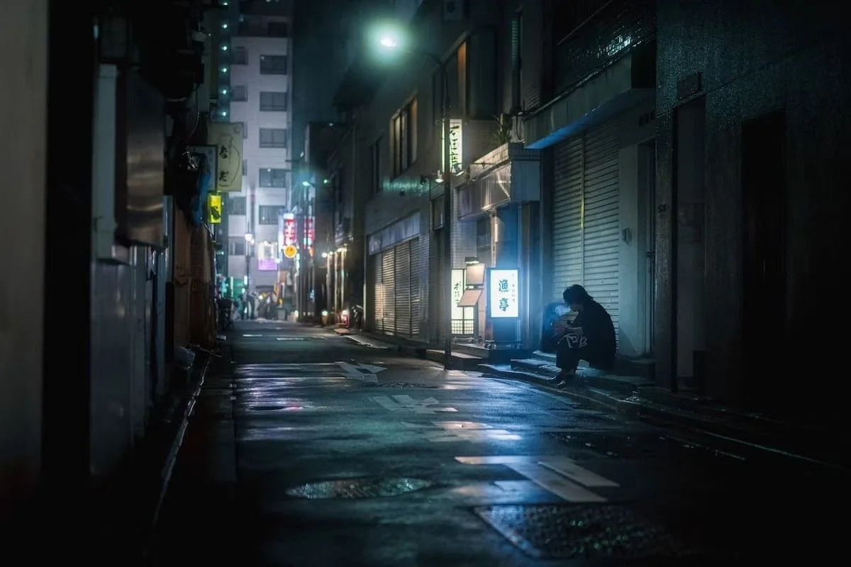 A person sitting alone on a dimly lit wet city street at night, illuminated by neon signs and streetlights, with closed shop storefronts surrounding. Tokyo alleys in Shimbashi.