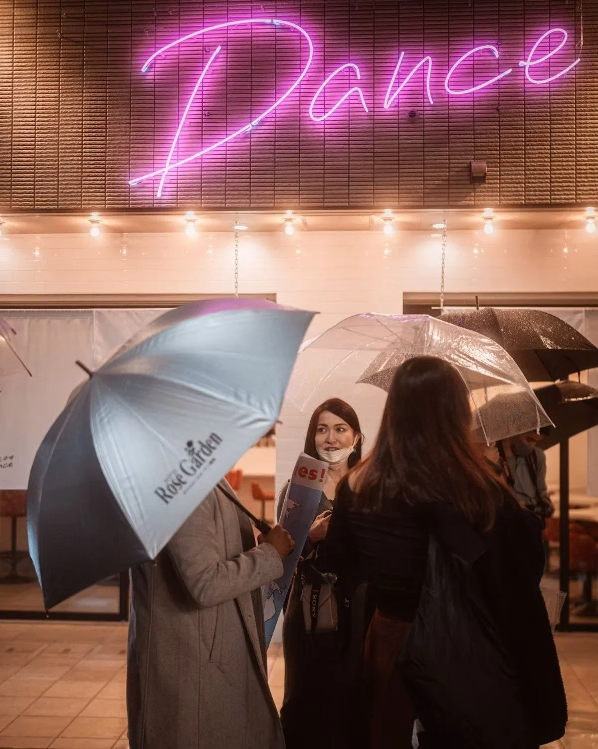 People standing outside in the rain with umbrellas, near a building with a neon sign that says 'Dance' in pink and purple lights shot in Shin Okubo Tokyo. 