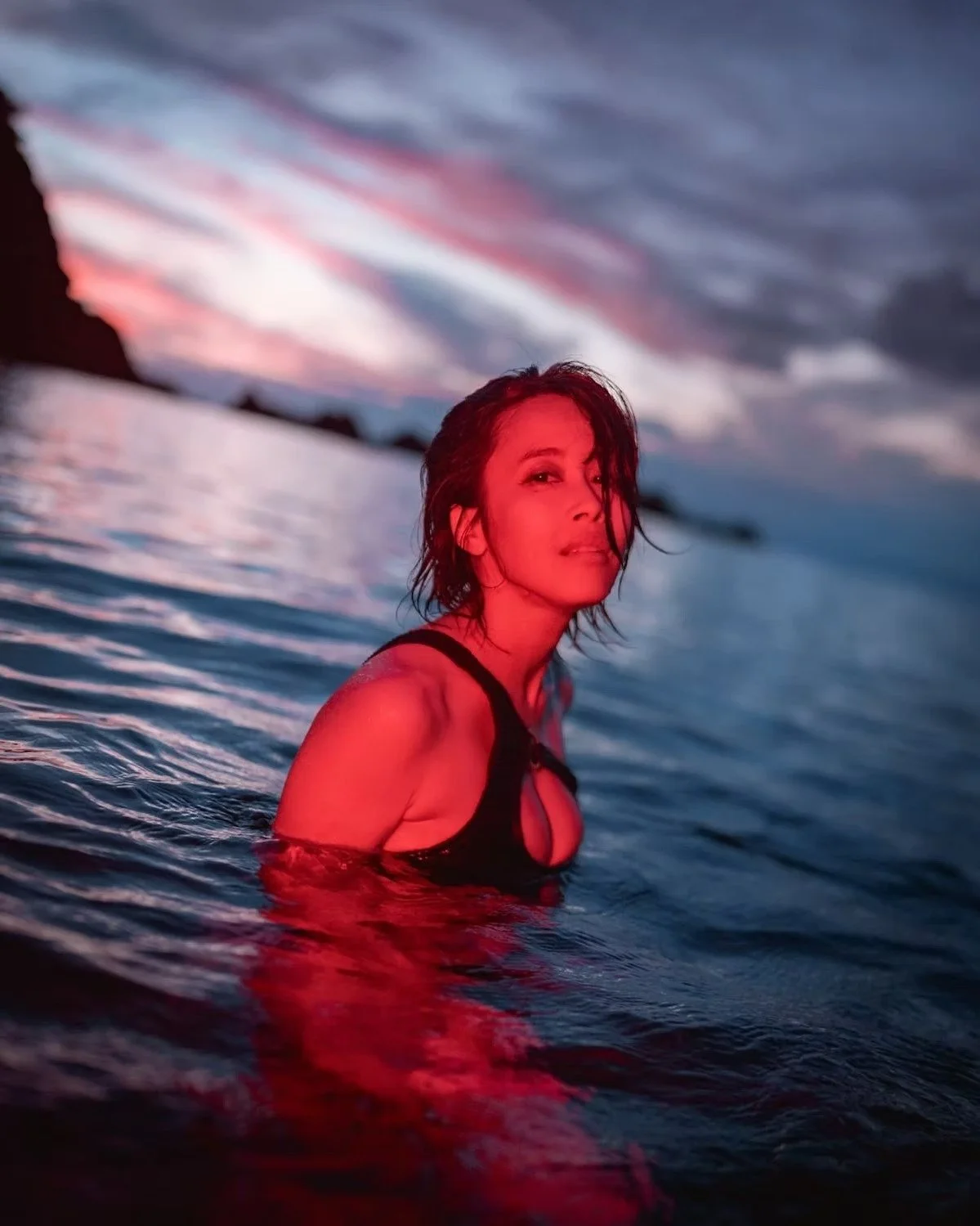A woman with wet hair in a black swimsuit wading in water during sunset with colorful sky reflections. Portrait photography by Tokyo photographer Deniz Demir.