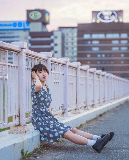 A girl in a floral dress sitting on the road with the Osaka skyline in the background, captured in a portrait.