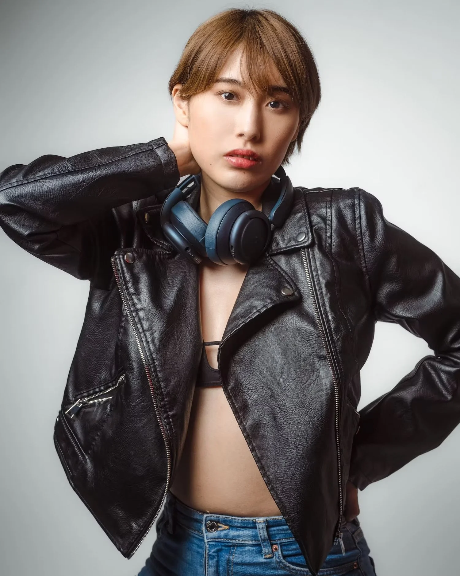 Person with short brown hair wearing a black leather jacket and blue jeans, with headphones around neck, posing against a plain gray background.