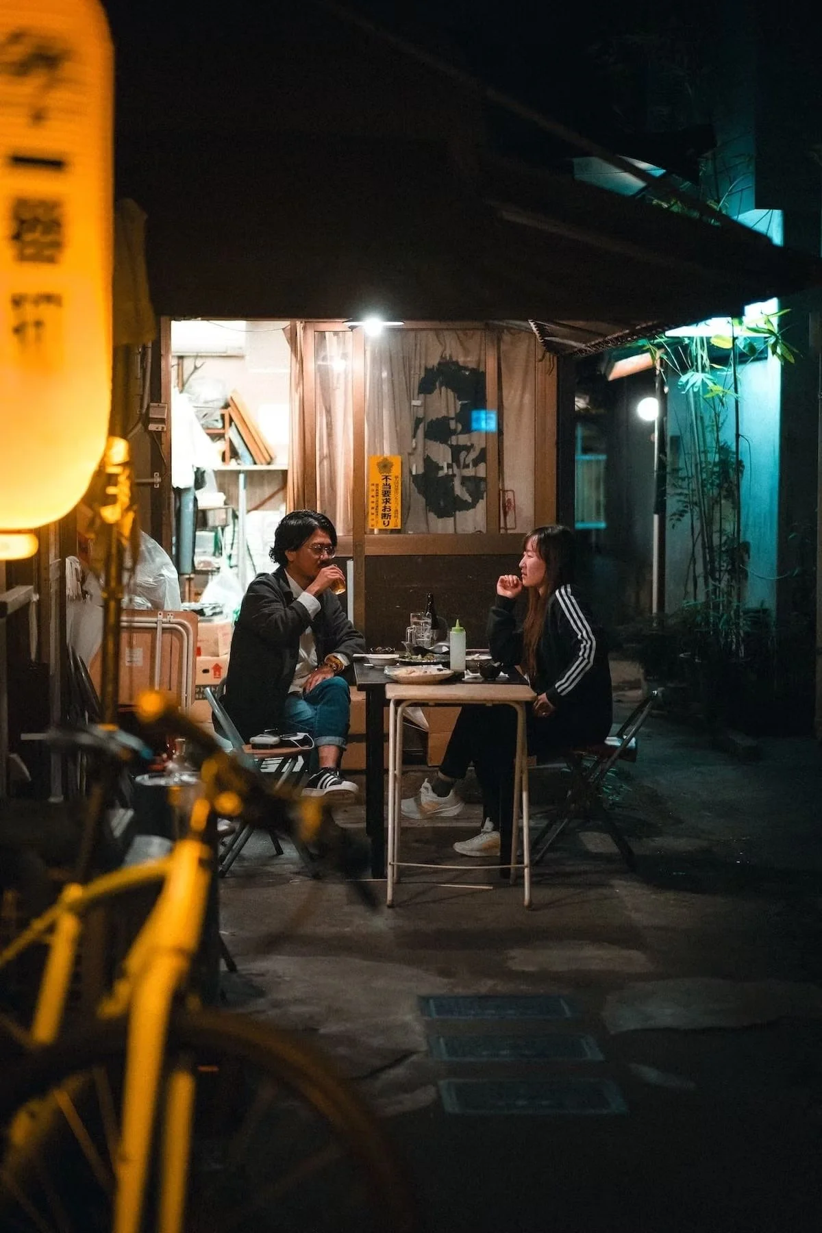 A man and a woman sit at a small outdoor table at night in Kagoshima Japan, having a meal together. There are drinks and food on the table, and they are engaging in conversation. The scene is lit softly, with a wooden wall, some plants, and hanging l
