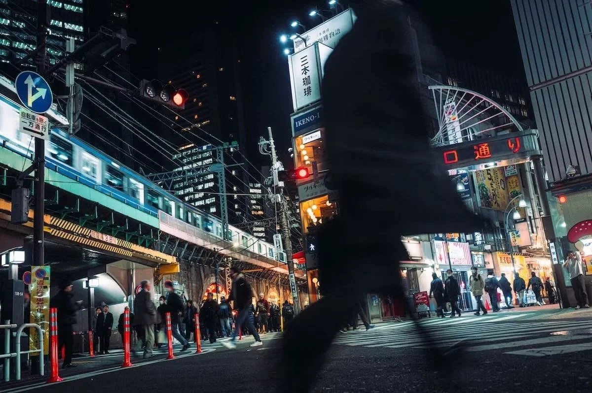 Nighttime street scene in Shimbashi Tokyo like a cyberpunk scene with pedestrians crossing the street, illuminated signs, and a train passing overhead on an elevated track, with tall buildings in the background.