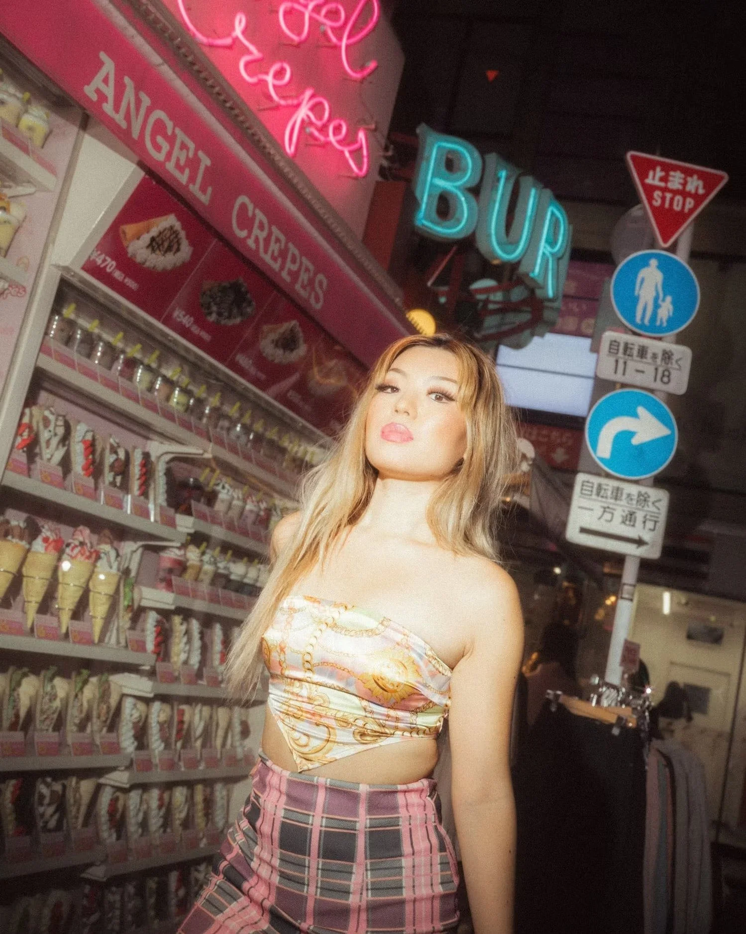 A young woman with long, wavy blonde hair posing in front of a store with neon signs for 'BUR' and 'LADY' and various signs and products, including ice cream cones, in the background.