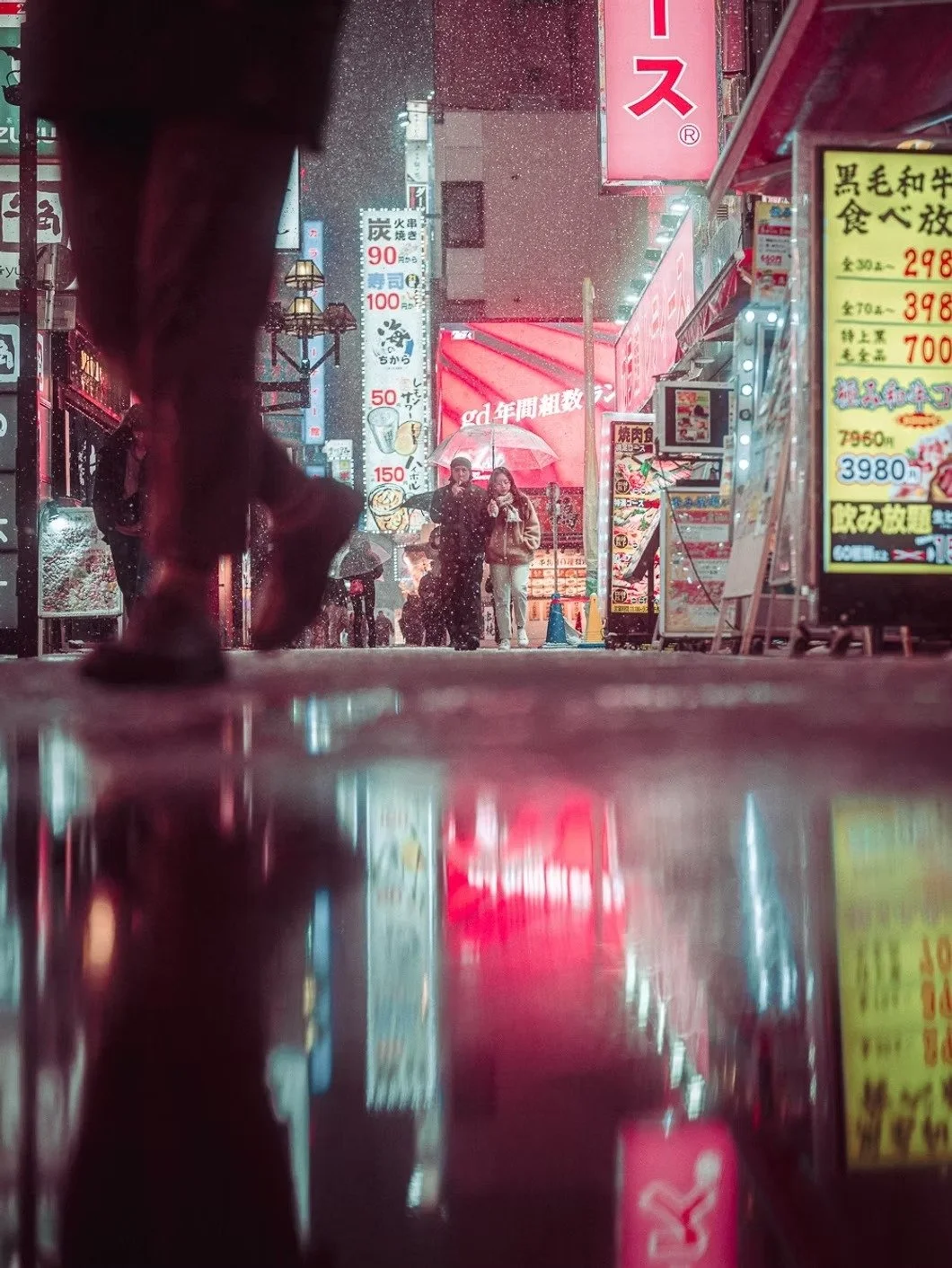 People walking on a neon-lit city street at night, some holding umbrellas in the rain, with various illuminated signs and advertisements in the background.