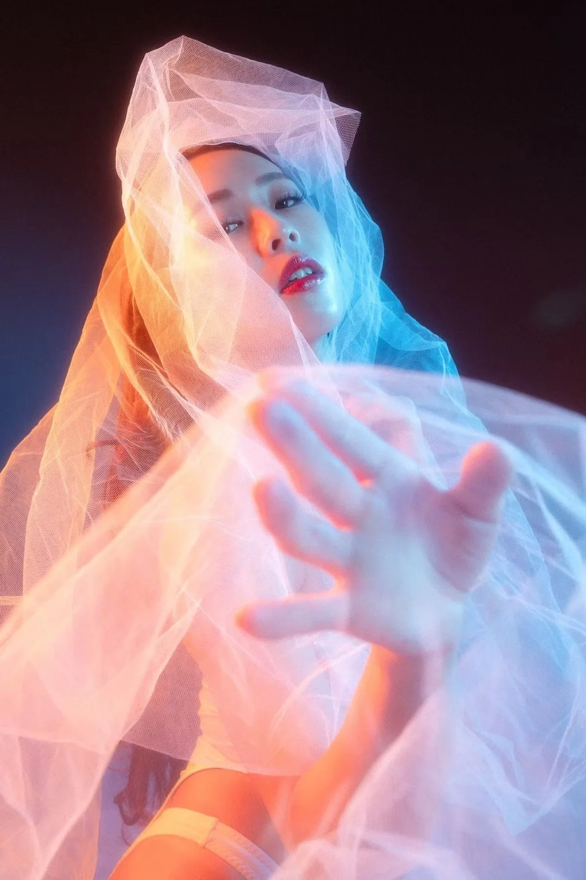 A woman with dark hair and makeup posing behind sheer pink and blue fabric, reaching forward with her hand.