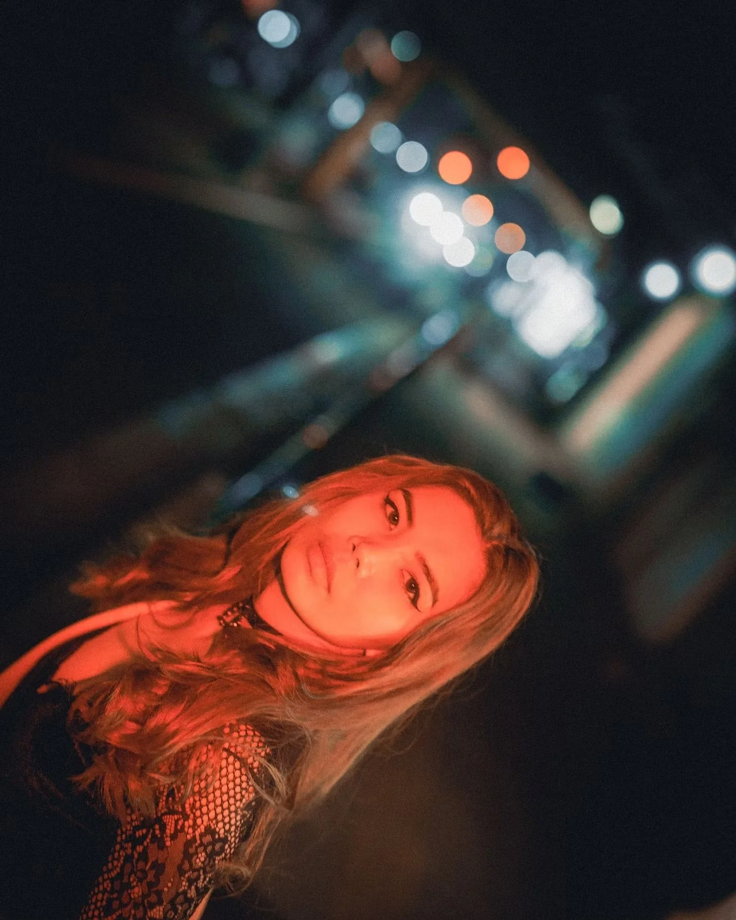 A woman with long wavy hair, wearing a black lace top, smiling and looking at the camera at night with colorful bokeh lights in the background.