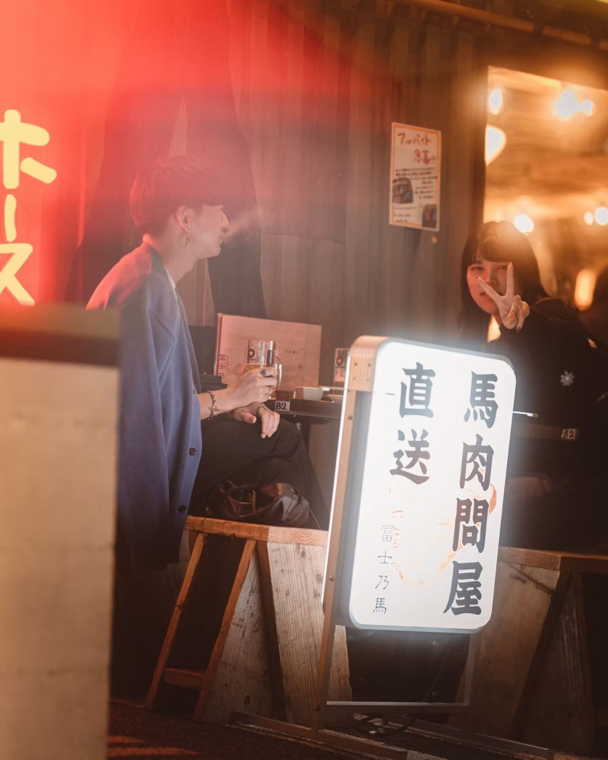 A Japanese couple sitting at a small restaurant table, one making a peace sign, with a bright signboard and warm lighting creating an inviting atmosphere. Shot by Tokyo street photographer Deniz Demir.