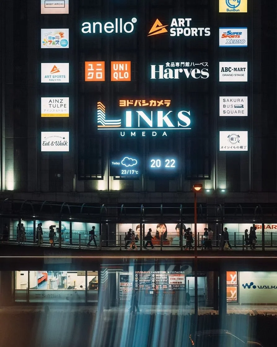 Nighttime city scene with large advertising screen displaying various brand logos and a weather forecast, with pedestrians crossing an elevated walkway below.