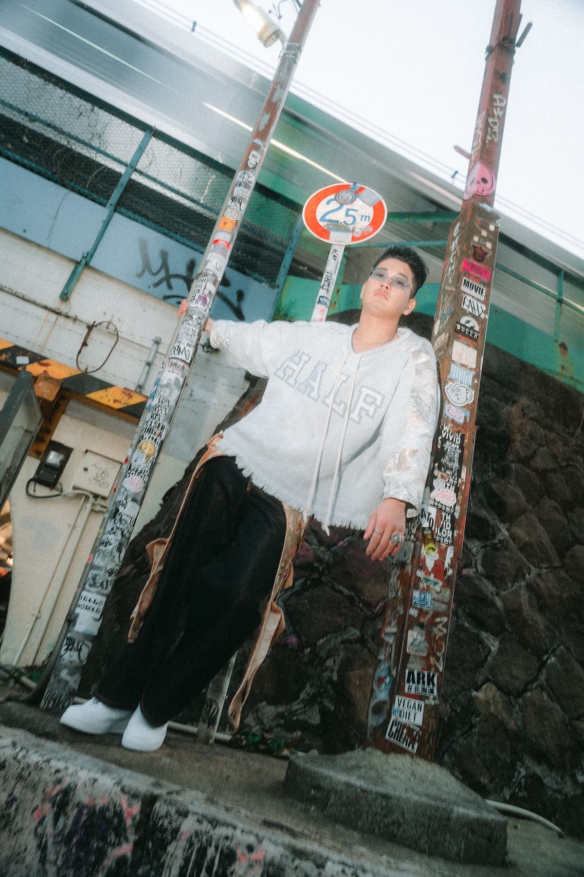 A person standing outdoors next to a graffiti-covered pole and a street sign indicating a height restriction of 2.5 meters. The individual is wearing glasses, a light-colored hoodie, black pants, and white shoes, with a backpack hanging from one shoulder, and has short hair styled upward. The background includes a stone wall and some graffiti on a fence.