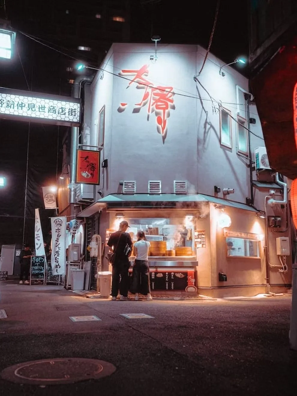 A nighttime street scene with a small restaurant or food stand emitting steam, with two people standing in front of it. The building has a large red Japanese kanji character on its white wall, illuminated by outside lights. There are signs and banners with Japanese writing, and the street appears to be in an urban area with other buildings around.