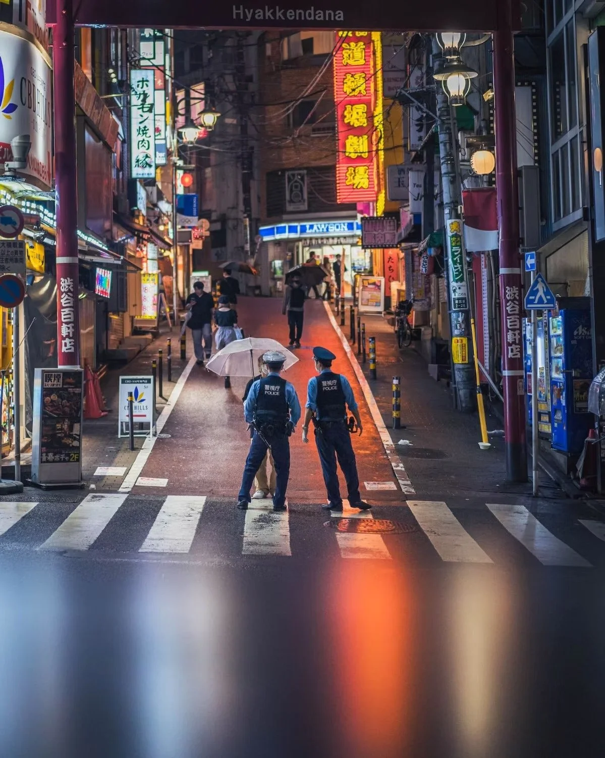 Two police officers are talking to a person on a pedestrian crosswalk on a rainy evening in a neon-lit city street in Japan. Several pedestrians are also walking with umbrellas. Night street photography in Shibuya by Deniz Demir.