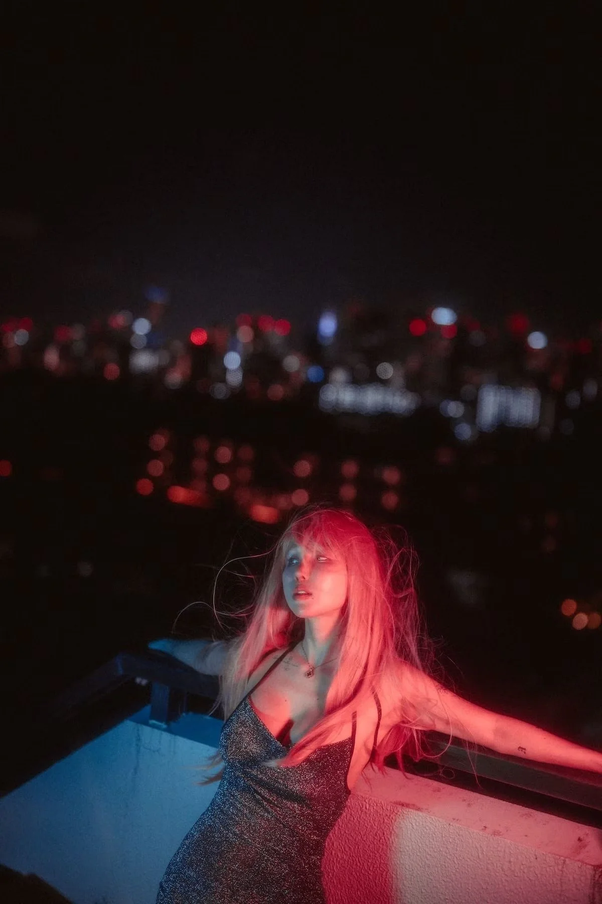 A woman with long blonde hair stands on a rooftop at night with a city skyline in the background. She is wearing a sparkly dress and is illuminated by pink and blue lighting.