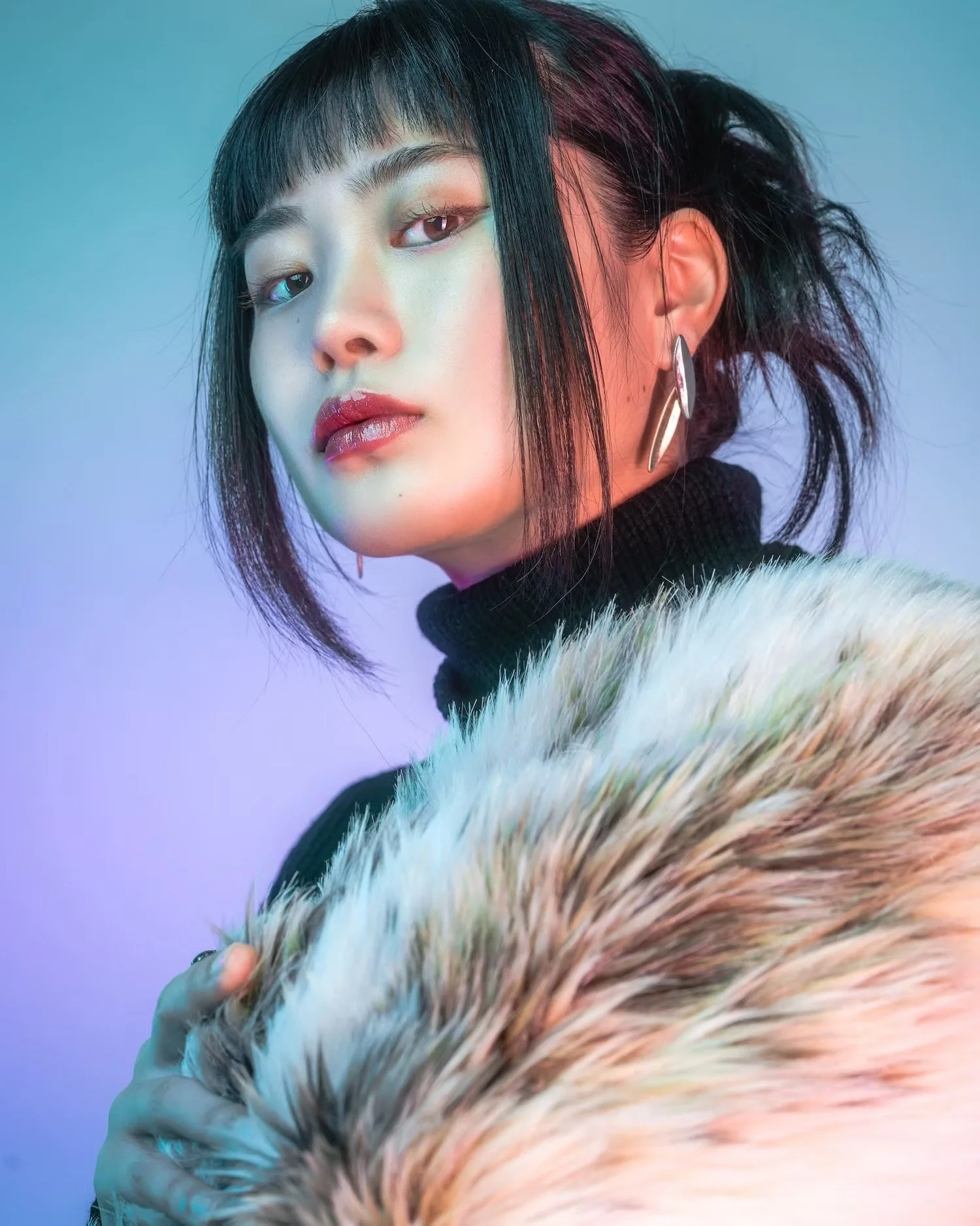 A woman with short black hair, wearing makeup, earrings, and a black turtleneck, is holding a large, multicolored fur.