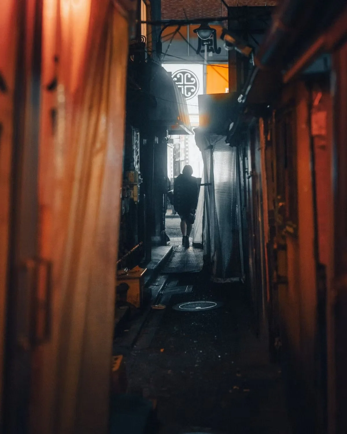 A hostess walking in a narrow alleyway, silhouetted against the bright street outside, with wooden walls and various objects lining the sides in Kabukicho Tokyo Japan.