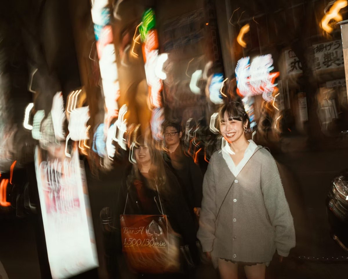A woman smiling at night outdoors with city lights blurred in the background in Shibuya using shutter drag.