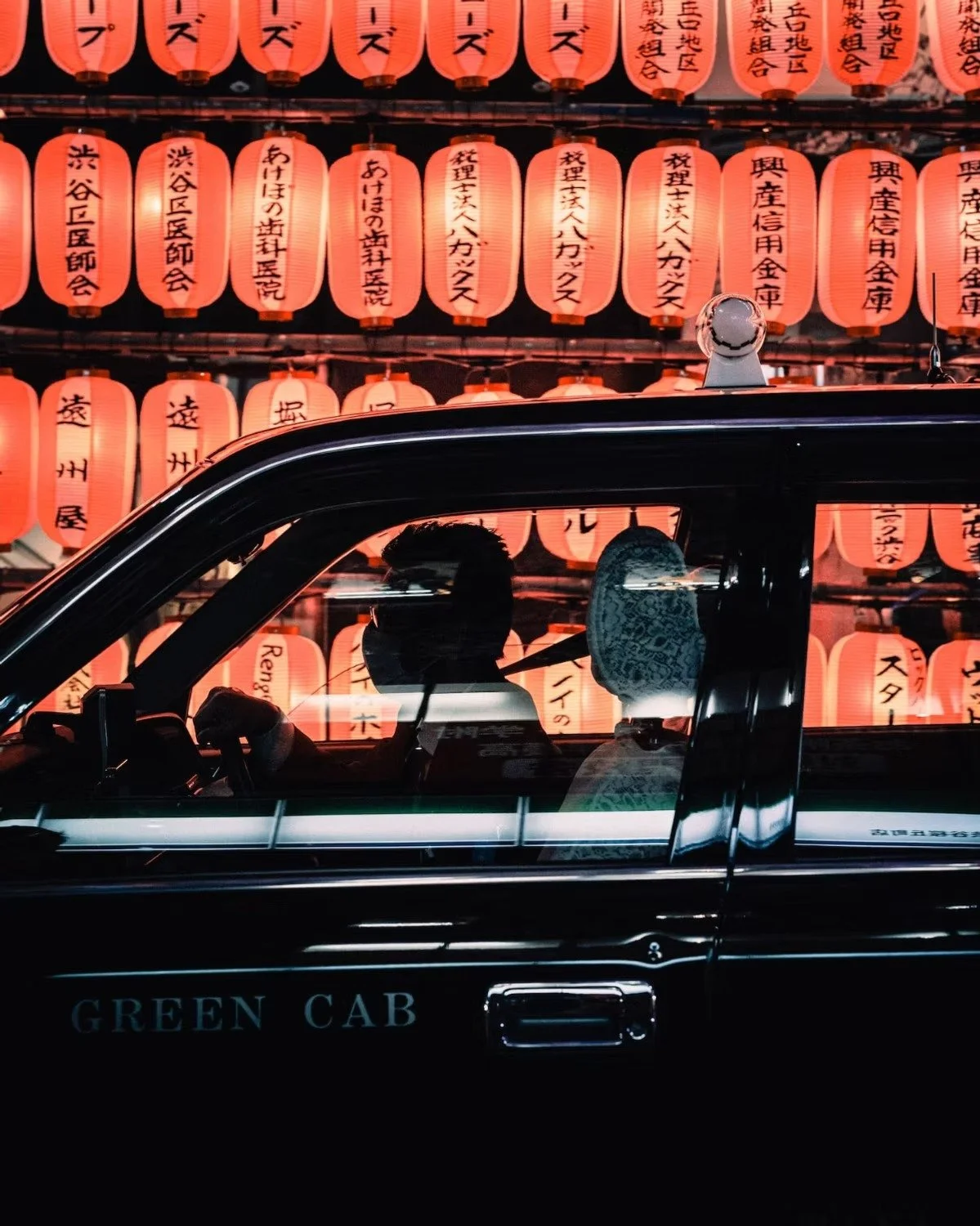 A black Tokyo taxi with two passengers inside, driving past a display of red Japanese paper lanterns with black writing, illuminated at night.