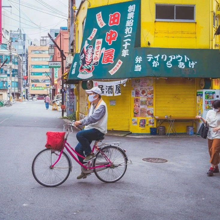 A cyclist riding down an empty street in front of a yellow building in Osaka, captured during slow travel.