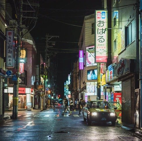 A dimly lit Tokyo street at night with neon signs reflecting on the wet pavement, as a car approaches and pedestrians walk in the distance.