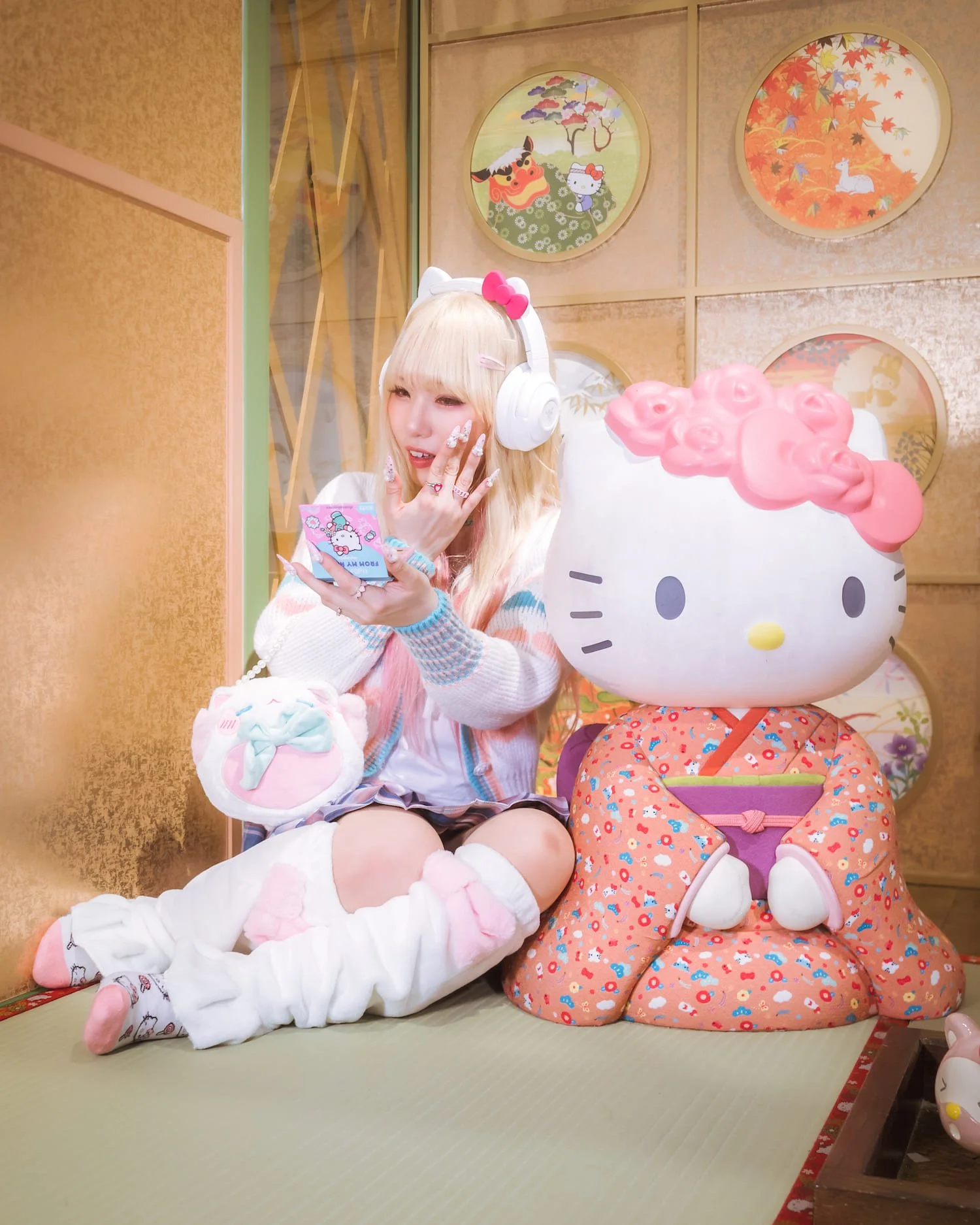 A woman with long blonde hair wearing large white Hello Kitty headphones, a pink bow, and a white outfit with pastel accents, sitting on a pink and white plush, holding a pink Hello Kitty themed cloth, sitting next to a large Hello Kitty plush with a pink curly hair, dressed in an orange kimono with a purple obi, in front of a golden wall with circular Japanese-style art.
