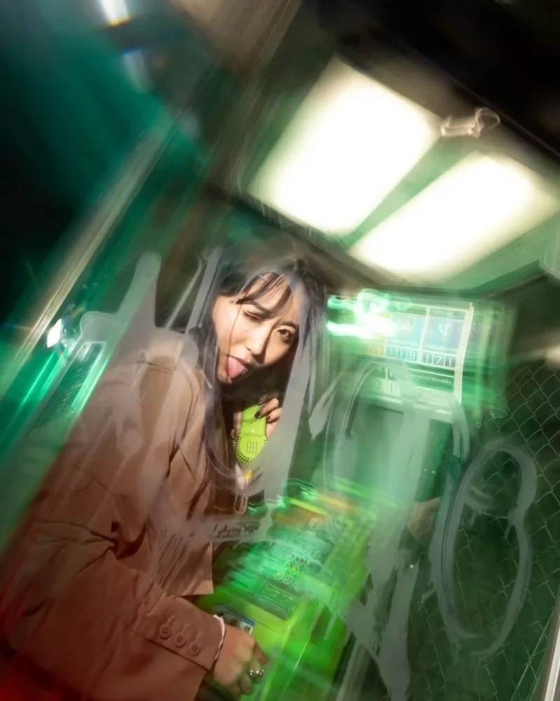 A woman with long hair and a beige trench coat posing playfully with her tongue out while holding a yellow phone inside a neon-green-lit area with arcade or gaming machines.