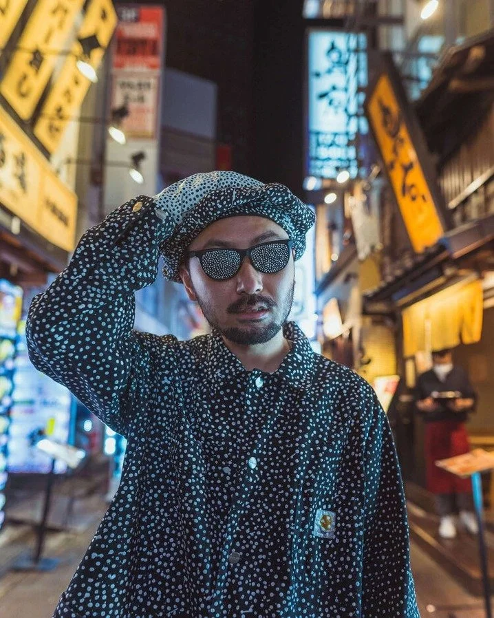 A man in a polka-dot outfit with sunglasses, standing in Kabukicho amidst colorful neon signs at night.