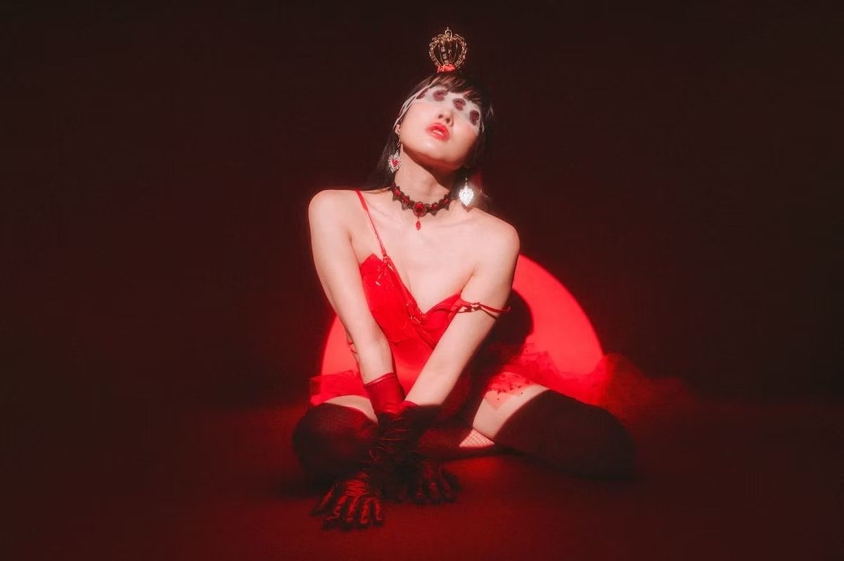 A woman dressed in red lingerie with gloves, wearing a crown and jewelry, sitting on the floor with a red circle behind her, against a dark background.