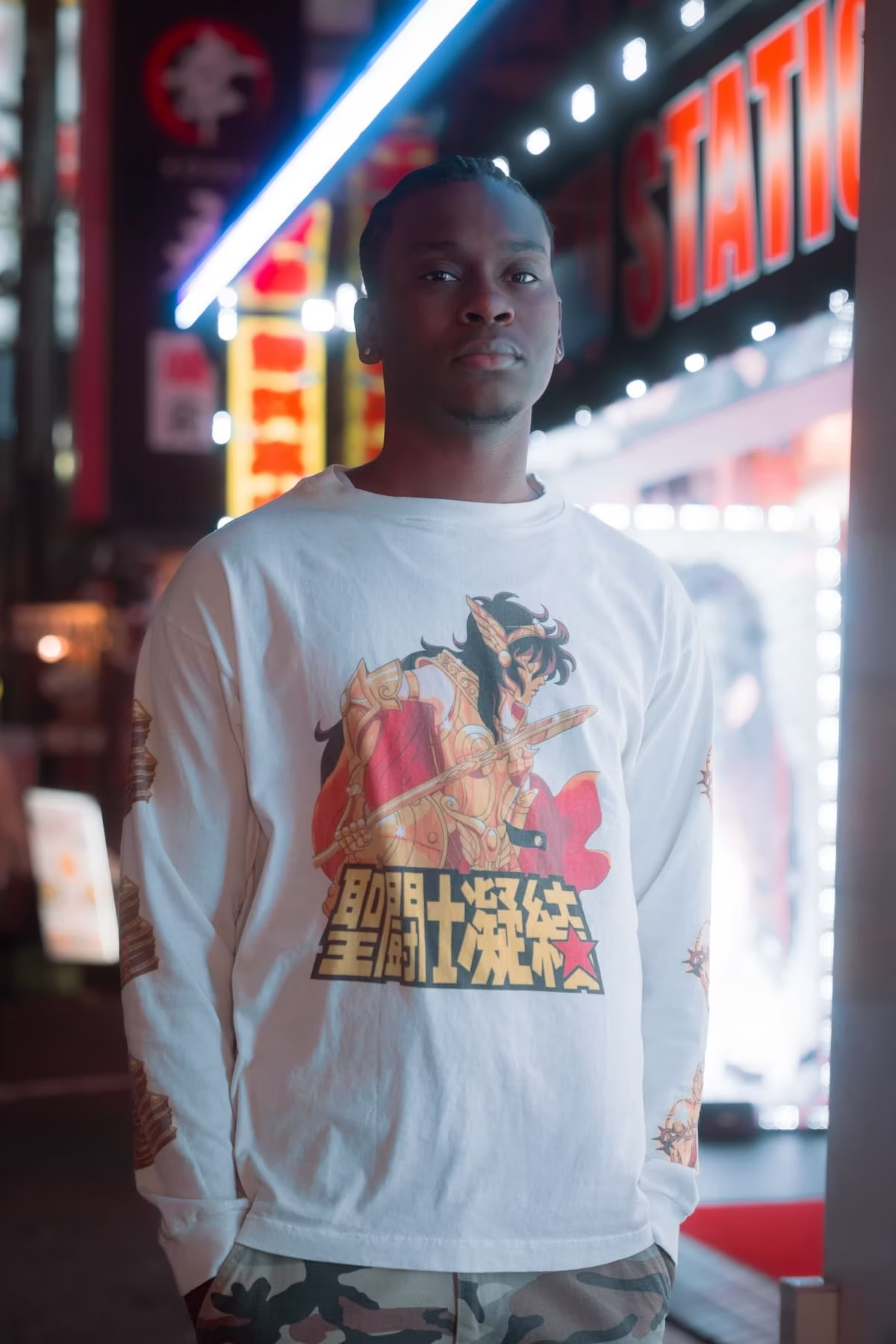 A man wearing a white long-sleeve shirt with a graphic print and camouflage pants stands on a brightly lit street in Tokyo, surrounded by neon lights and signs.